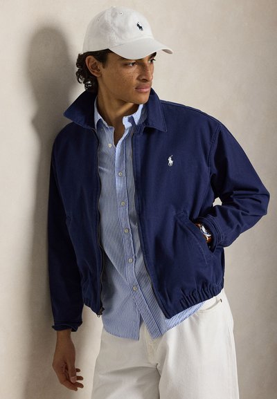 Homme portant une casquette blanche, une veste bleu marine sur une chemise rayée bleue, et un pantalon blanc, regardant vers la droite avec une main dans la poche de la veste.