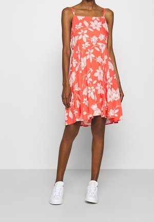 Nainen, joka pukeutunut hihoittomaan korallinväriseen mekkoon, jossa on valkoinen kukkakuosi, ja valkoiset tennarit, seisoo yksinkertaisen vaalean taustan edessä.