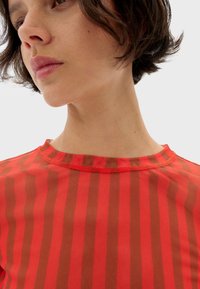 Rød og brunstribet top med rund halsudskæring. Stoffet virker let og transparent med kontrasterende syningsdetaljer langs kanten.