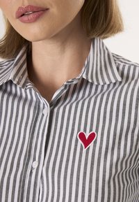 Šedobílý pruhovaný košile s červeným vyšívaným srdcem na levé straně, s klasickým límcem a zapínáním na knoflíky.