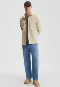 Beige corduroy overhemd met knopen, groen T-shirt eronder, lichtblauwe jeans en tan schoenen. Een pet maakt de casual outfit compleet.