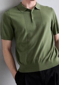 Hombre vestido con una camiseta de polo de punto de manga corta en verde oliva, con paneles verticales texturizados, combinada con pantalones negros, de pie contra una pared gris claro.