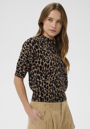 Femeie cu păr lung și castaniu, purtând un top cu mânecă scurtă și imprimeu leopard și pantaloni bej cu talie înaltă, cu mâna în buzunar, privind spre stânga.