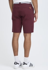 Shorts bordeaux en tissu texturé, avec un patch de marque, des poches plates et une ceinture à motifs, associés à des baskets blanches.