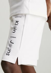 Witte katoenen shorts met zwarte tekstafdrukken met de tekst "Klein" en "super Jeans." Voorzien van zijsplitten en een relaxed fit ontwerp.