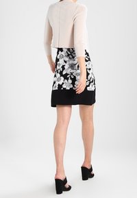 Femme portant une jupe fleurie noir et blanc, un cardigan court rose pâle, et des chaussures à talons ouverts noires, se tenant sur un fond blanc.