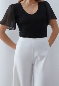 Černý top s průsvitnými nařasenými rukávy a výstřihem do V, doplněný vysokým bílými kalhotami. Hladká textura, střih na míru.