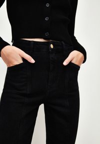 Jeans de tiro alto negros hechos de mezclilla, con un botón de metal dorado, bolsillos laterales y una parte superior negra con textura de canalé con cierre de botones.