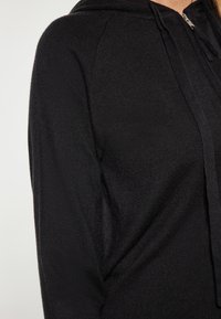 Černá mikina na zip s měkkou tkaninou, raglánovými rukávy a texturovaným povrchem. Obsahuje kapuci se šňůrkou a přední zip.