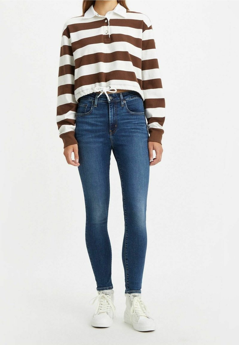 Femme portant un polo court à manches longues rayé marron et blanc, jean skinny bleu taille haute et baskets blanches montantes.