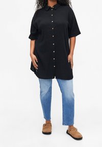 Chemise noire à manches courtes et coupe décontractée, avec des boutons dorés et un tissu texturé. Portée avec un jeans droit bleu clair et des chaussures brunes.