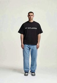 Jovem em pé, vestindo uma t-shirt oversized preta com o texto "2Y STUDIOS", jeans azul claro e ténis preto e branco, em fundo liso.