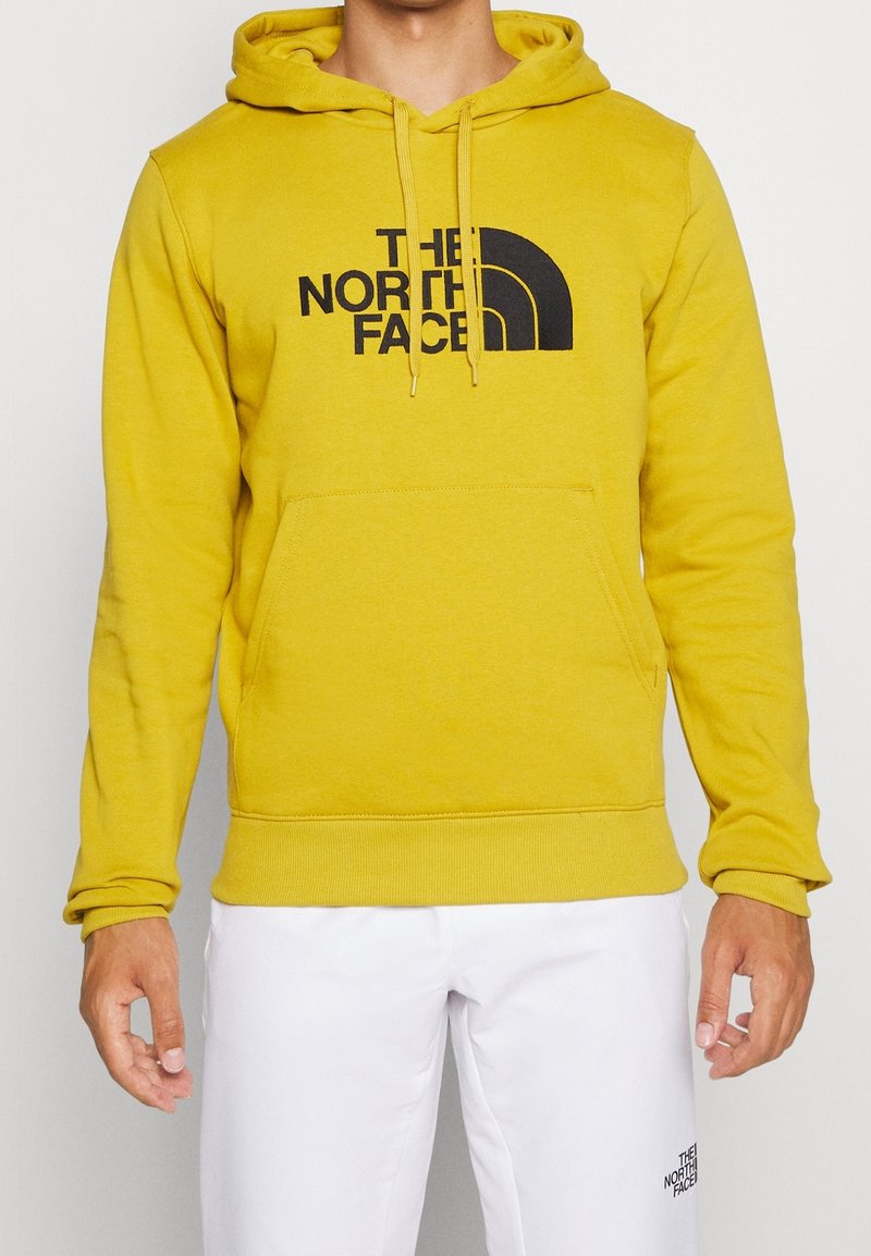 Homme portant un sweat à capuche jaune moutarde The North Face et un pantalon blanc avec le logo The North Face sur la cuisse gauche, debout devant un fond uni.