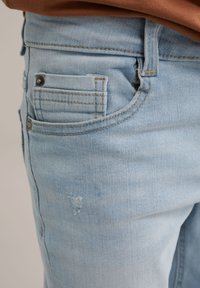 Gai zila džinsu bikses ar nolietotu plāksteri, kontrastējošām šuvēm un aizmugurējo kabatu detaļu. Materiāls ir mīksts un nedaudz nēsāts.