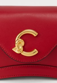 Κόκκινη δερμάτινη τσάντα με εμφανείς ραφές και χρυσό μεταλλικό κούμπωμα σε σχήμα στυλιζαρισμένου γράμματος C με διακοσμητικό στοιχείο που ενώνεται.