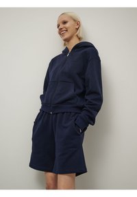 Námornícky modrá mikina na zips s kapucňou, prednými vreckami a rebrovanými manžetami, skombinovaná s nohavicami na kolená v rovnakom odtieni, vyrobená z jemného materiálu.
