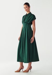 Žalia medvilninė suknelė su apykakle, trumpomis pūstomis rankovėmis ir prigludusia juosmeniu, turinti sklandų, raukinį sijoną. Derinama su rudais raštais papuoštais aukštakulniais.