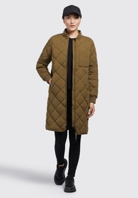 Manteau long matelassé vert olive avec col montant, fermeture éclair et poches latérales, associé à des leggings noirs et des baskets.