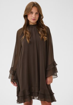 Jeune femme aux longs cheveux ondulés porte une robe ample, marron foncé, à plusieurs couches avec des manches longues transparentes et un ourlet à volants, debout devant un fond uni.