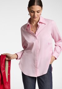 Maglietta a maniche lunghe rosa chiaro con colletto abbottonato, decorata con un piccolo logo di fiore ricamato. Abbinata a jeans blu scuro e una giacca rossa.