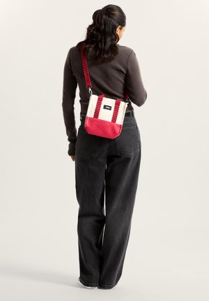 Kabelka přes tělo v krémové a červené barvě, s texturovaným popruhem, obdélníkovým tvarem a logem na nášivce. Materiál vypadá odolně a hladce.