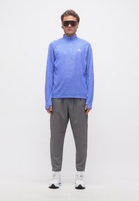 Νέος άνδρας που φορά μπλε αθλητική μπλούζα με μακριά μανίκια, γκρι παντελόνια, λευκά αθλητικά παπούτσια και μεγάλα κόκκινα γυαλιά ηλίου, στέκεται μπροστά σε απλό φόντο.