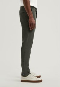 Slim-fit olijfgroene broek met een gestructureerde afwerking en zijnaaddetails, gedragen met witte sneakers op een neutrale achtergrond.