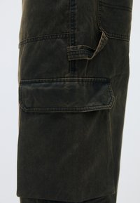 Σκούρο ελαιόλαδο cargo παντελόνι με υφή, που διαθέτει μια μεγάλη πλαϊνή τσέπη και ανθεκτικές ραφές. Σχεδιασμένο για πρακτικότητα και αντοχή.