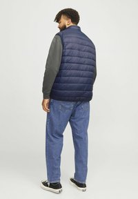 Marineblaues Steppweste mit horizontalen Nähten, hohem Kragen und glatter Textur, über einem grauen Langarmshirt und blauen Jeans getragen.