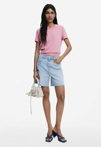Camiseta ajustada de color rosa claro, shorts de mezclilla azul claro con bolsillos y sandalias de tacón azules. La modelo sostiene un bolso pequeño blanco.