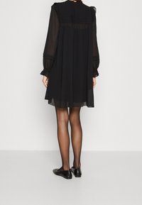 Robe en chiffon noire à manches longues, avec des poignets volantés et des détails plissés à l'arrière. Portée avec des ballerines noires et des collants transparents.