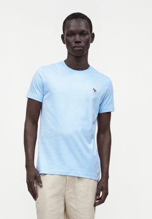 Bărbat purtând un tricou cu mâneci scurte bleu cu un logo mic de zebră pe piept și pantaloni bej, stând în fața unui fundal simplu.