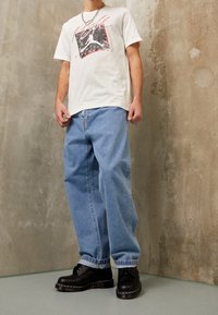 Camiseta gráfica blanca con diseño en rojo y negro, jeans holgados de mezclilla azul claro y zapatos negros con cordones y suela gruesa.