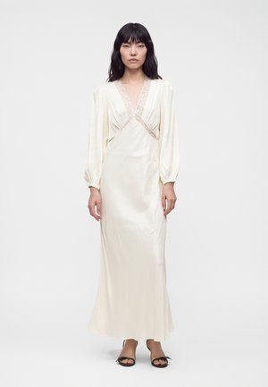 Femme portant une longue robe en satin crème avec bordure en dentelle et manches longues, debout sur un fond blanc uni.