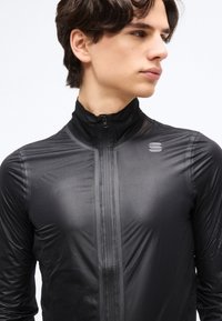 Νεαρός άνδρας που φοράει ένα μαύρο αθλητικό σακάκι με φερμουάρ, κοιτάζοντας προς τα πλάγια, με κοντά σκούρα μαλλιά, μπροστά σε απλό λευκό φόντο.