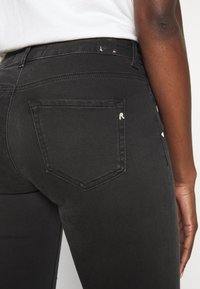 Mustat denimi-farkut, joissa on istuva malli, takataskussa tunnistettava ompelukuosi ja sileä pinta. Räätälöity leikkaus korostaa muotoa.