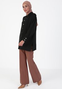 Black long-sleeve blouse with gold buttons, side slits, paired with brown wide-leg trousers and beige heels. Smooth fabric texture.