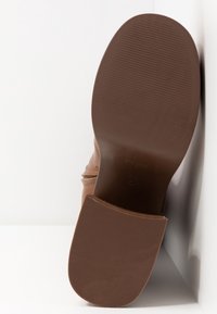 Semelle d'une chaussure en cuir marron avec un talon en caoutchouc texturé et l'avant-pied lisse, photographiée sur un fond blanc.