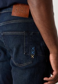 Jeans din denim închis, cu o aplicație din piele texturată etichetată "Scotch & Soda". Accente brodate în albastru și galben pe buzunarul din spate.