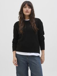 Μαύρο πλεκτό πουλόβερ με υφή, στρογγυλή λαιμόκοψη και μακριά μανίκια, φορεμένο πάνω από ένα λευκό πουκάμισο, συνδυασμένο με φαρδιά μπλε τζιν.