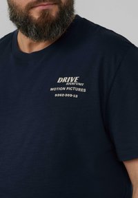 Granatowa bawełniana koszulka z okrągłym dekoltem. Z przodu znajduje się biały nadrukowany tekst: "DRIVE STUNT UNIT MOTION PICTURES 9362-309-13".