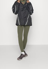 Czarna lekka kurtka z odblaskowymi akcentami, zielone legginsy oraz ciemnoszare buty sportowe z jasnoszarymi podeszwami i białe skarpetki.