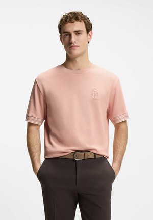 Homme aux cheveux bouclés portant une chemise rose à manches courtes et un pantalon marron, debout les mains dans les poches devant un fond uni.