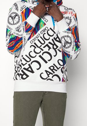 Weißer Hoodie mit großem schwarzem "Carlo Colucci"-Schriftzug und mehrfarbigen abstrakten Mustern auf den Ärmeln, getragen von einer Person, die den Kragen anpasst.