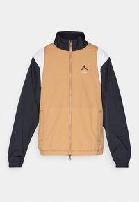 Tuulitakki, jossa on vaaleanruskea runko, mustat hihat ja valkoiset yksityiskohdat. Takissa on pystypääntie, vetoketjullinen suljenta ja ikoninen logo rinnassa.