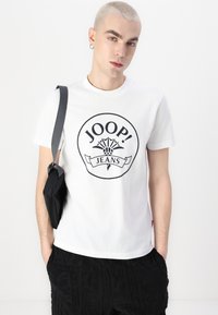 Bílá bavlněná trička se zaobleným černým logem s nápisem "JOOP! JEANS." Model má na sobě černé texturované kalhoty a nese černou tašku.