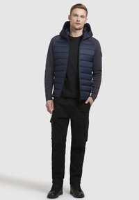 Veste rembourrée bleu marine avec capuche, manches grises et fermeture éclair à l'avant, associée à un pantalon cargo noir et des bottes noires. Design simple et fonctionnel.