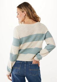 Frau mit welligem blonden Haar, die silberne Kreolen trägt, einen cremefarbenen und hellblauen gestreiften Pullover sowie Blue Jeans anhat, und mit dem Rücken zur Kamera steht.