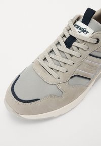 Svijetlo siva tenisica s kombinacijom antilopa i mrežastih materijala, s naglascima u mornarsko plavoj boji, okruglim nosom i klasičnim dizajnom s vezicama. Vidljiv Wrangler brand.