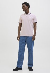 Lyserød polo-shirt med sort krave og kant, parret med blå denimjeans. Modellen står med hænderne i lommerne og har sorte sko på.